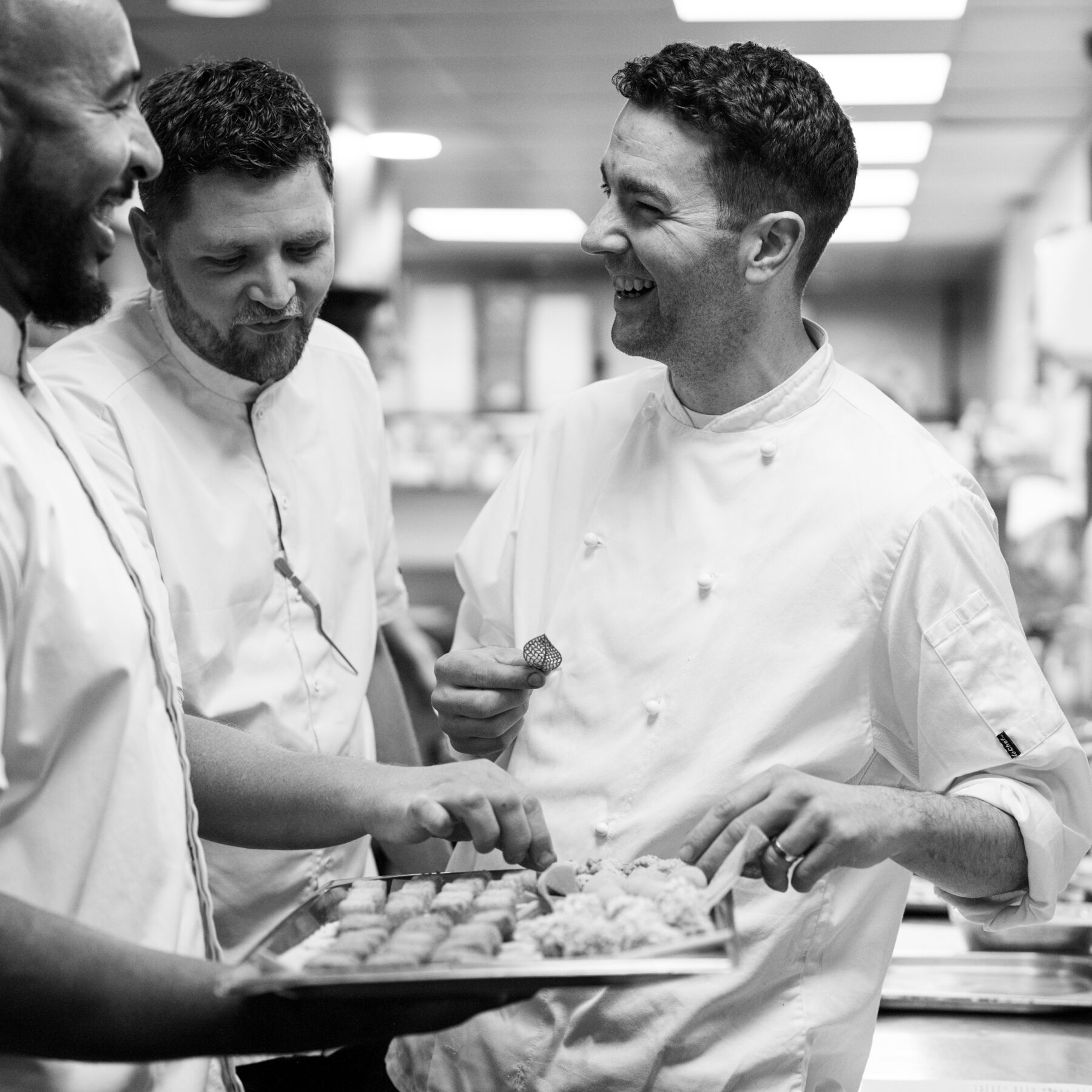 A black and white photo of three chefs in the kitchen smiling whilst holding a tray of food